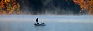 Person fishing alone on a small boat in a misty lake, surrounded by autumn trees and calm water.