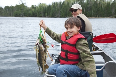 A boy in a red life jacket holds up several fish in a canoe, with an adult sitting behind him on a lake.