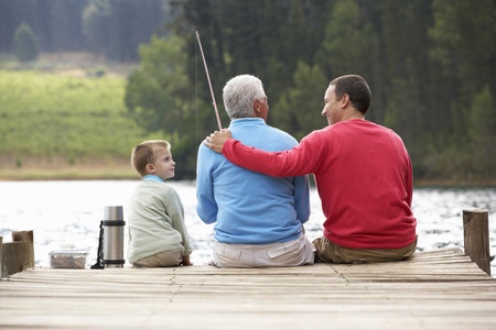 Three people sit on a wooden dock by a lake, with one holding a fishing rod and another with an arm around the third.