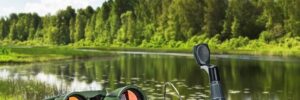 Fishing equipment, binoculars, and bait on a wooden surface near a calm lake with trees and a blue sky in the background.