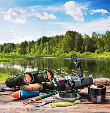 Fishing equipment, binoculars, and bait on a wooden surface near a calm lake with trees and a blue sky in the background.