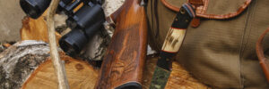 A shotgun, binoculars, a knife, and a canvas bag rest on a cut tree stump with stacked firewood in the background.