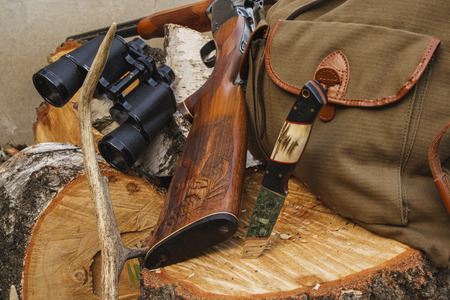 A shotgun, binoculars, a knife, and a canvas bag rest on a cut tree stump with stacked firewood in the background.