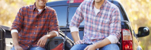 Two men in plaid shirts sit on the tailgate of a pickup truck with fishing gear on a sunny day.