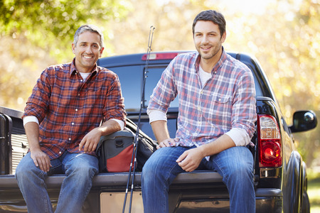 Two men in plaid shirts sit on the tailgate of a pickup truck with fishing gear on a sunny day.