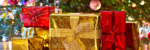 Several wrapped gifts in gold and red paper sit on a wooden table in front of a decorated Christmas tree.