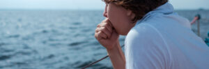 A person in a white shirt leans on a railing and looks thoughtfully at the water on a boat.