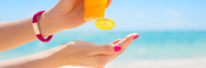 Person squeezing sunscreen from a yellow bottle onto their hand at a beach with blue sky and ocean in the background.