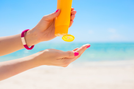 Person squeezing sunscreen from a yellow bottle onto their hand at a beach with blue sky and ocean in the background.