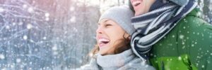 Two people dressed in winter clothing smile and laugh together outdoors as snow falls in a snowy, wooded area.