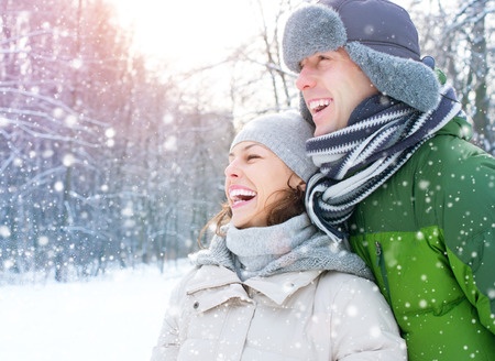 Two people dressed in winter clothing smile and laugh together outdoors as snow falls in a snowy, wooded area.