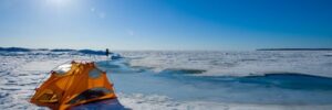 An orange tent is set up on a snowy, frozen landscape under a clear blue sky with bright sunlight.