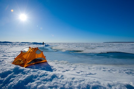 An orange tent is set up on a snowy, frozen landscape under a clear blue sky with bright sunlight.