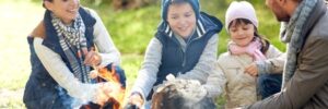 A family of four sits around a campfire outdoors, roasting marshmallows and smiling together.