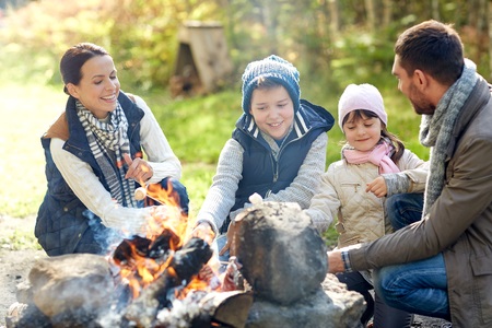 A family of four sits around a campfire outdoors, roasting marshmallows and smiling together.