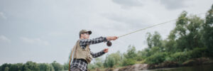 Person fly fishing while standing in shallow river water, surrounded by trees under a cloudy sky.