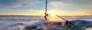 Ice auger, fishing rod, and ice hole on a frozen lake at sunset with snow and ice in the foreground.