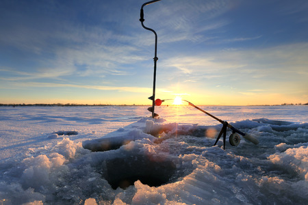 Ice auger, fishing rod, and ice hole on a frozen lake at sunset with snow and ice in the foreground.