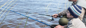 An adult and a child sit by a riverbank, fishing with rods on a cloudy day.