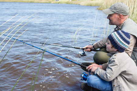 An adult and a child sit by a riverbank, fishing with rods on a cloudy day.