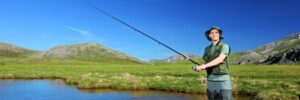 Person standing in shallow water with a fishing rod, wearing outdoor gear, with grassy hills and blue sky in the background.