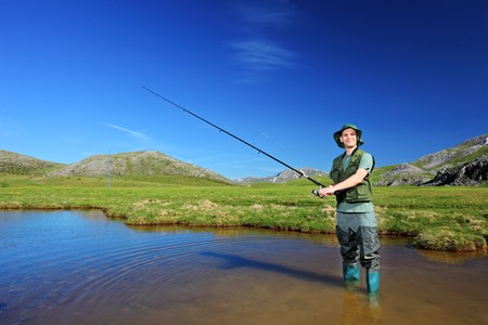 Person standing in shallow water with a fishing rod, wearing outdoor gear, with grassy hills and blue sky in the background.