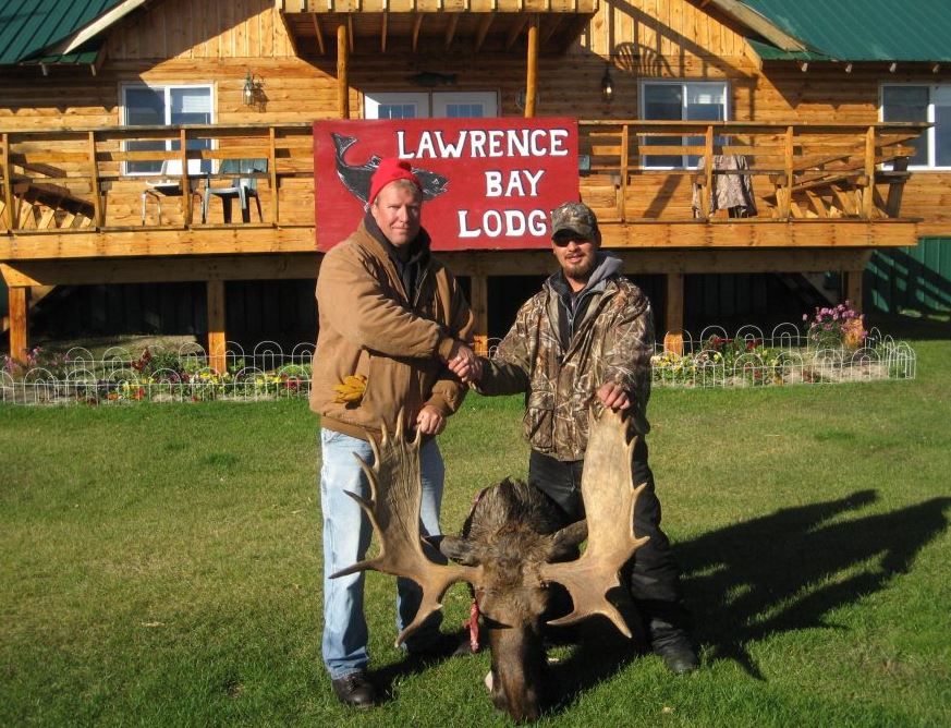 Two men shake hands in front of Lawrence Bay Lodge, posing with a large moose rack on the grass.