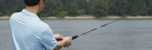 A man in a light blue shirt is fishing by a body of water with trees visible in the background.