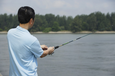 A man in a light blue shirt is fishing by a body of water with trees visible in the background.