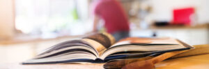Open cookbook on a wooden kitchen counter with wooden spoons; person blurred in the background.