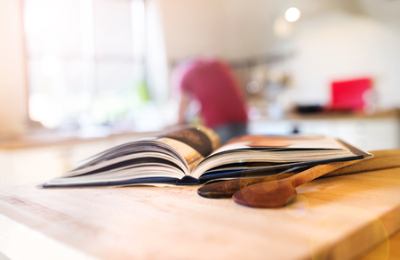 Open cookbook on a wooden kitchen counter with wooden spoons; person blurred in the background.