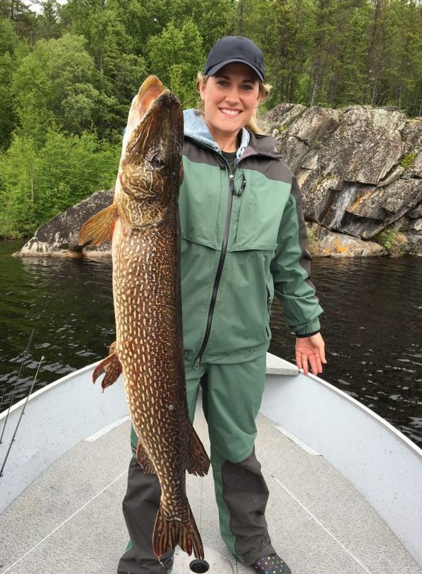 Person standing on a boat holding a large fish with a rocky, forested shoreline in the background.