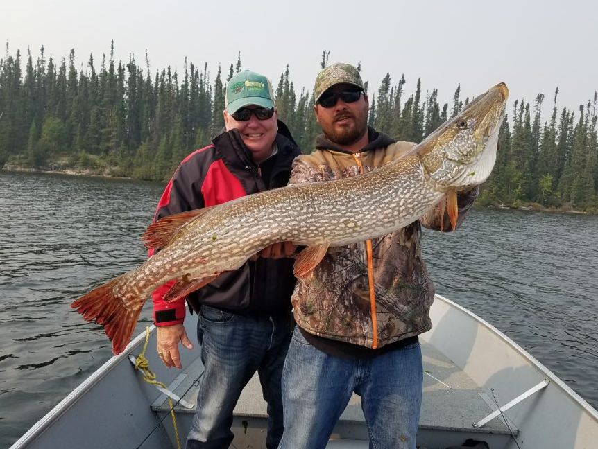 Two men in a boat hold a large, freshly caught fish with a forest and lake in the background.