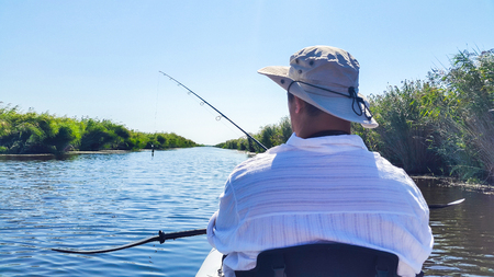 Person wearing a hat sits in a boat, fishing on a calm river surrounded by greenery under a clear blue sky.