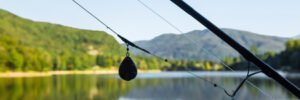 Close-up of a fishing rod with a sinker by a calm lake, with green hills and trees in the background.