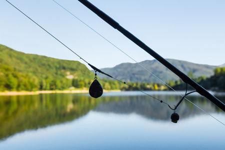 Close-up of a fishing rod with a sinker by a calm lake, with green hills and trees in the background.