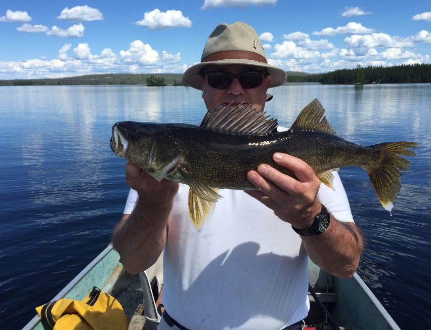 Person in a hat and sunglasses holding a large fish on a boat, with a lake and forested shoreline in the background.