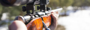 Person aiming a bolt-action rifle with a scope outdoors, background blurred with trees and snow.