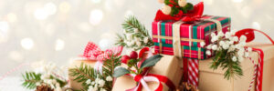 Three wrapped gifts decorated with ribbons, pine cones, and greenery on a white surface with a blurred festive background.