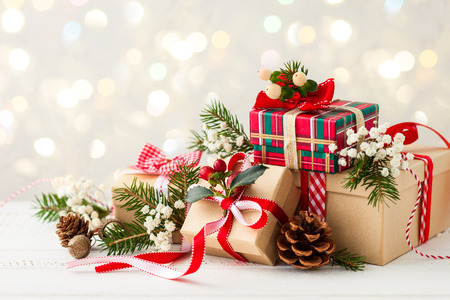 Three wrapped gifts decorated with ribbons, pine cones, and greenery on a white surface with a blurred festive background.