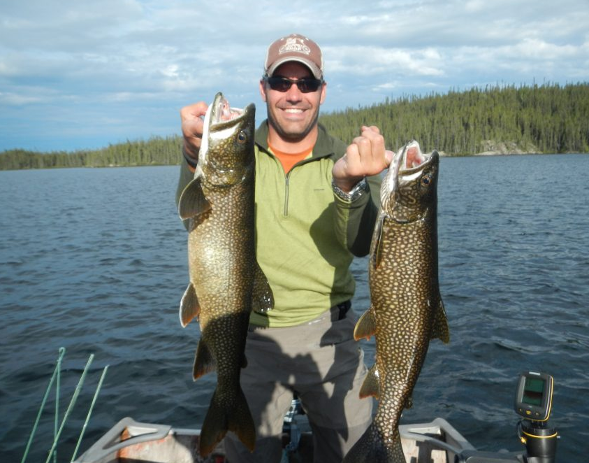 Man in sunglasses and a hat holds two large fish while standing on a boat in a lake with forest in the background.