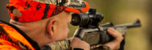 Person in camouflage and orange hunting gear aiming a rifle with a scope outdoors.