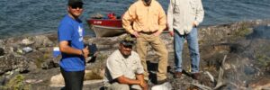 Four men by a lake with a boat in the background, cooking food over an open fire on the rocky shore.