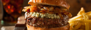 A cheeseburger with caramelized onions and blue cheese on a bun, served with French fries on a wooden board.