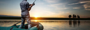 Person standing in an inflatable boat fishing at sunset on a calm lake with a clear sky and distant trees.