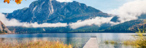 Wooden dock extends into a calm lake with mountains and autumn trees in the background under a blue sky.