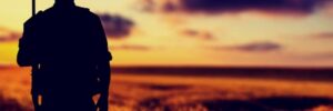Silhouette of a person with a rifle standing in a field at sunset, with clouds in the sky.