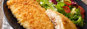 Breaded fish fillet served with lemon slices and a fresh mixed salad on a black plate.