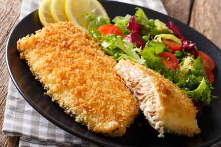 Breaded fish fillet served with lemon slices and a fresh mixed salad on a black plate.