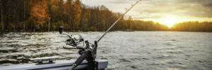 A fishing rod is mounted on a boat over a lake, with autumn trees and a sunset in the background.